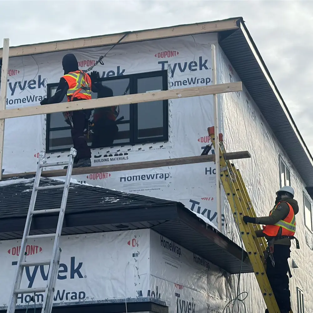 Two construction workers, one on a ladder and the other on a platform, install siding on a second-story window of a house under construction wrapped.