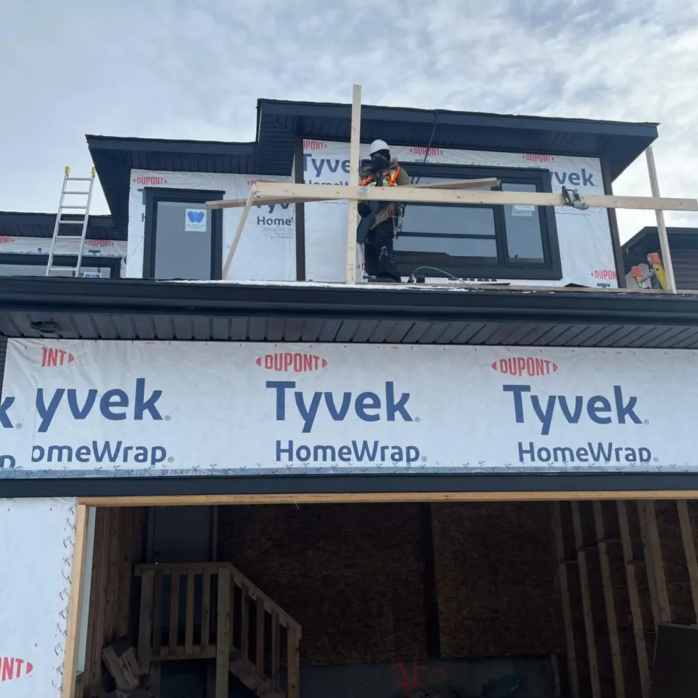Alt Text: A construction worker on a second-story platform installs exterior siding on a house with a ladder leaning against the structure.